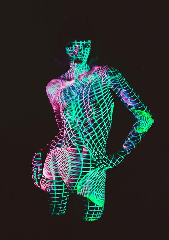 A woman is covered in projected neon green and pink grid patterns against a dark background.