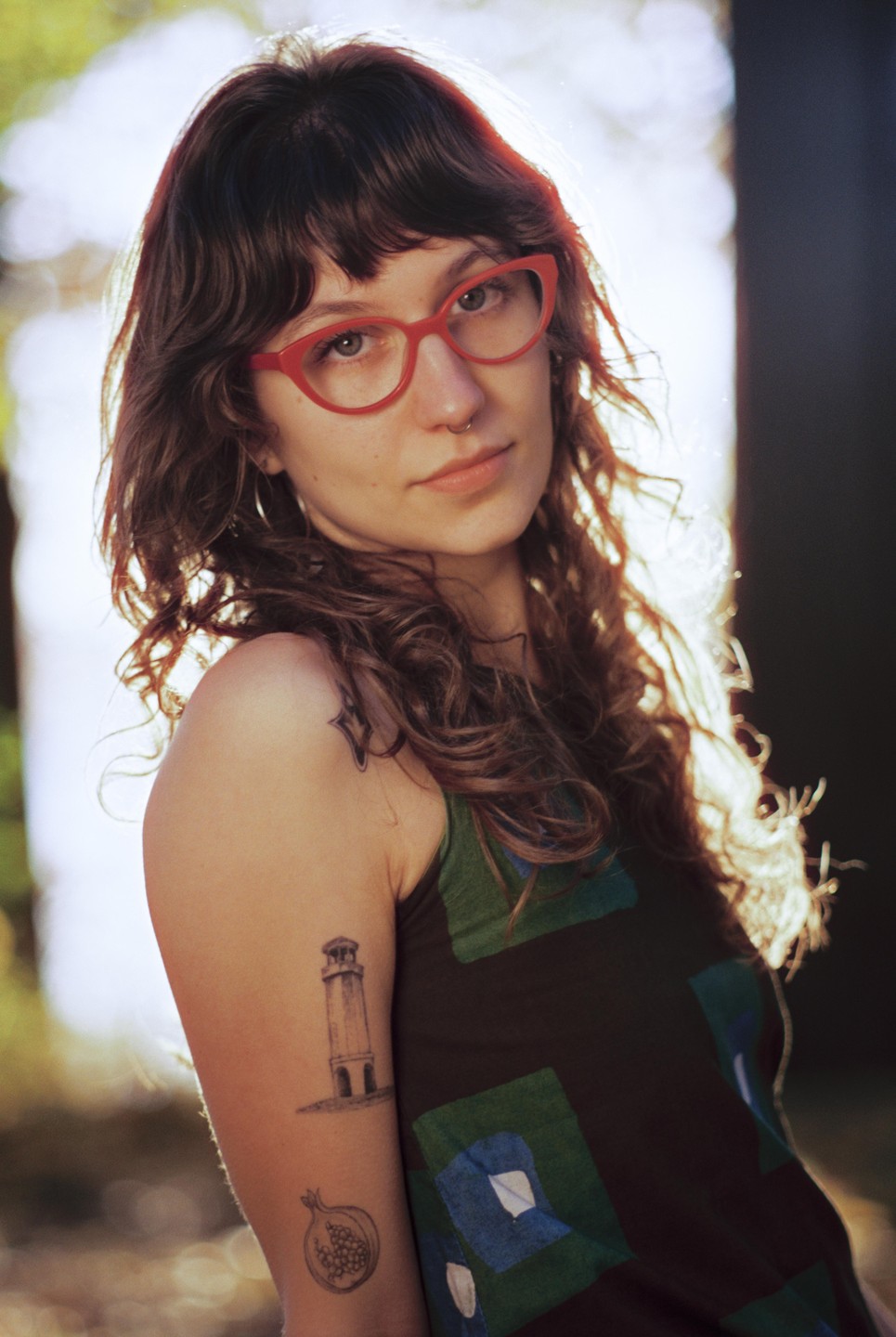 A woman with long, curly hair and red glasses looks directly at the viewer, with tattoos visible on her arm.
