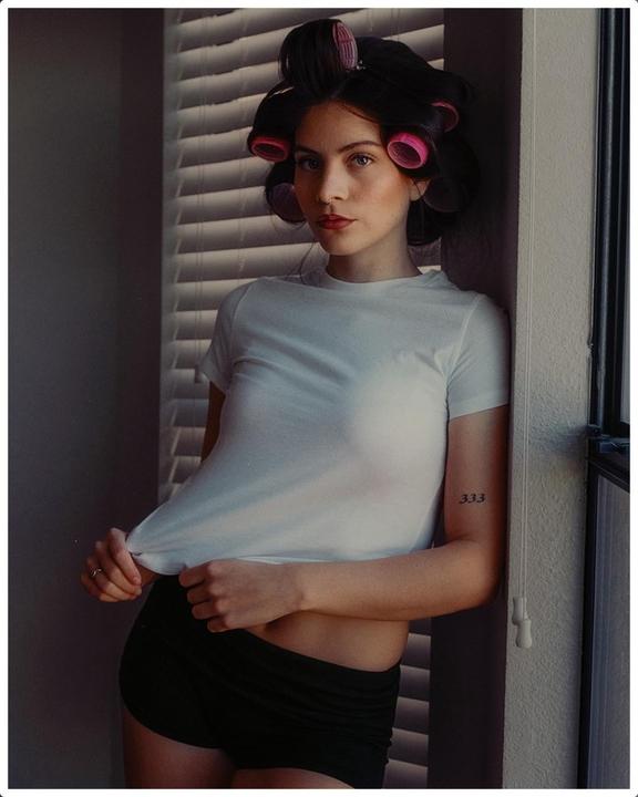 A woman with hair rollers poses in a white t-shirt and black shorts next to a window with blinds.