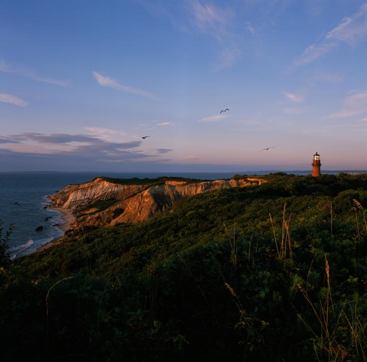 A lighthouse stands on a cliff overlooking the ocean at sunset, with birds flying in the sky.