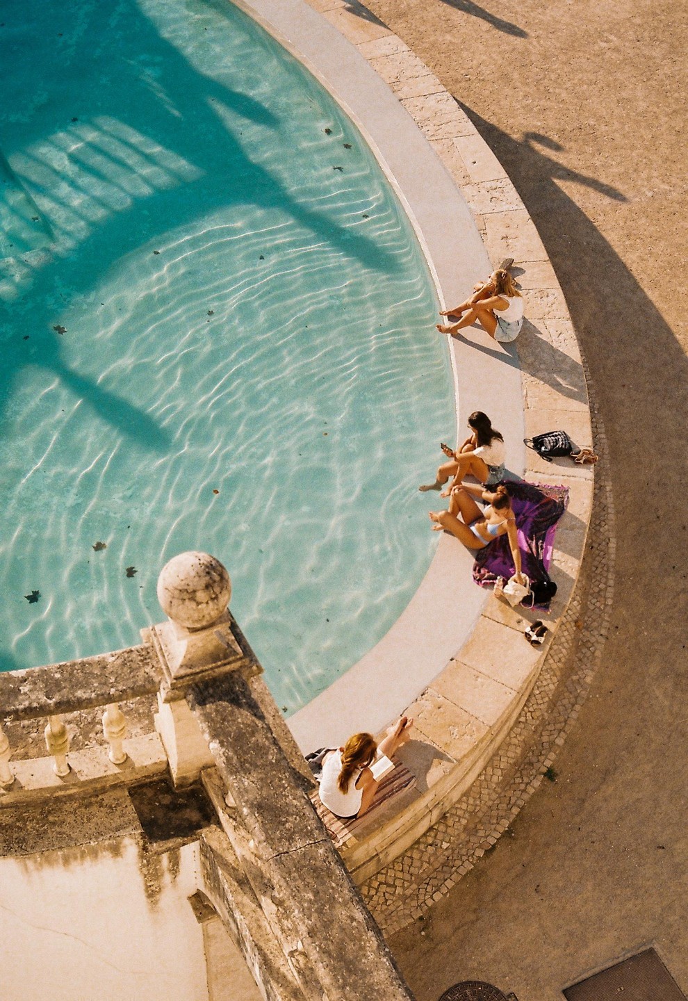 Four women relax by the edge of a pool with clear blue water and shadows cast across it.