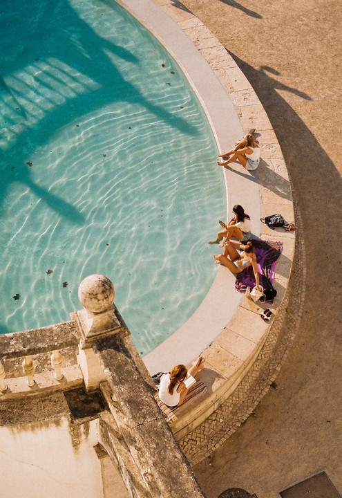 Four women relax by the edge of a pool with clear blue water and shadows cast across it.