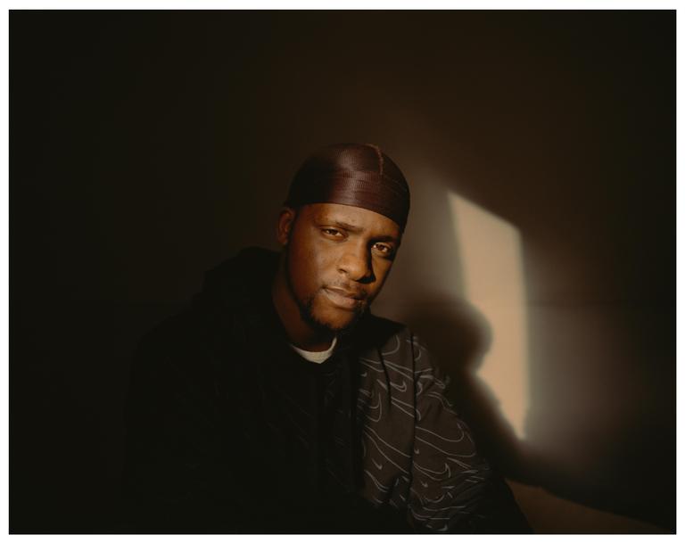A man wearing a durag and a patterned shirt is illuminated by a shaft of sunlight.