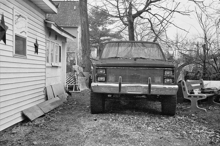 A black truck is parked on a gravel driveway next to a house with bare trees in the background.