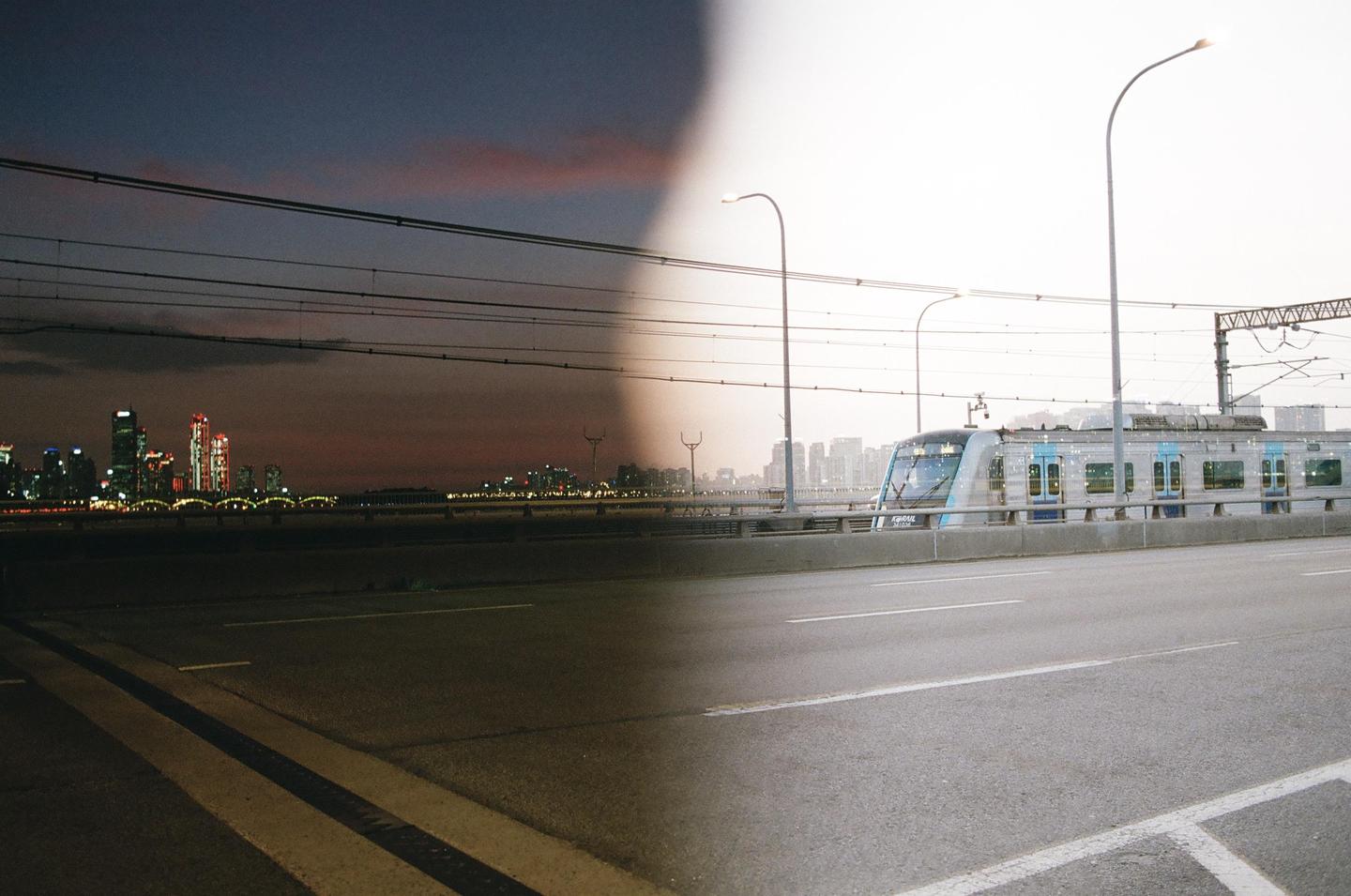 A double exposure shows a train on a bridge in front of a city skyline at night and during the day.