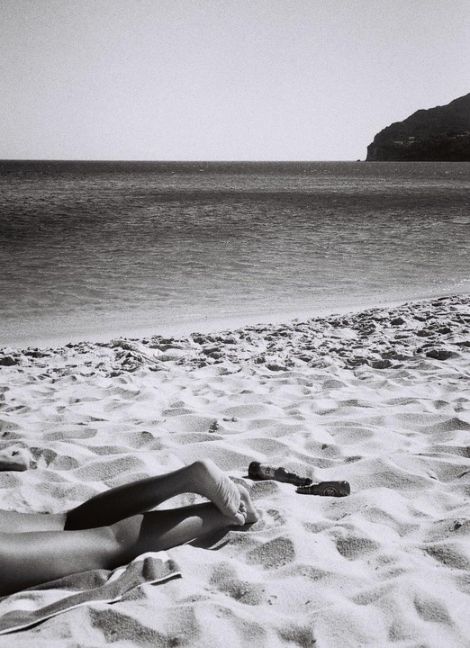 A person's legs and feet are visible on a sandy beach next to two bottles, with the ocean and a distant mountain in the background.