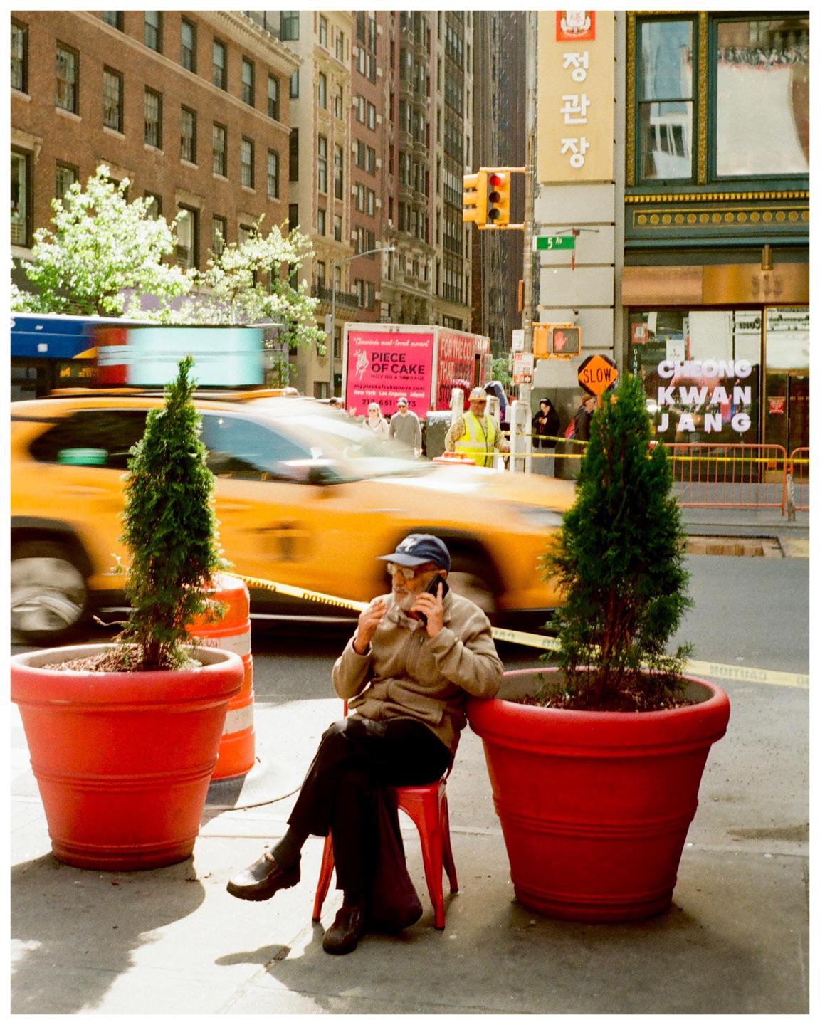 A man sits on a red chair on a city street, talking on a phone, with a yellow taxi cab speeding past in the background.