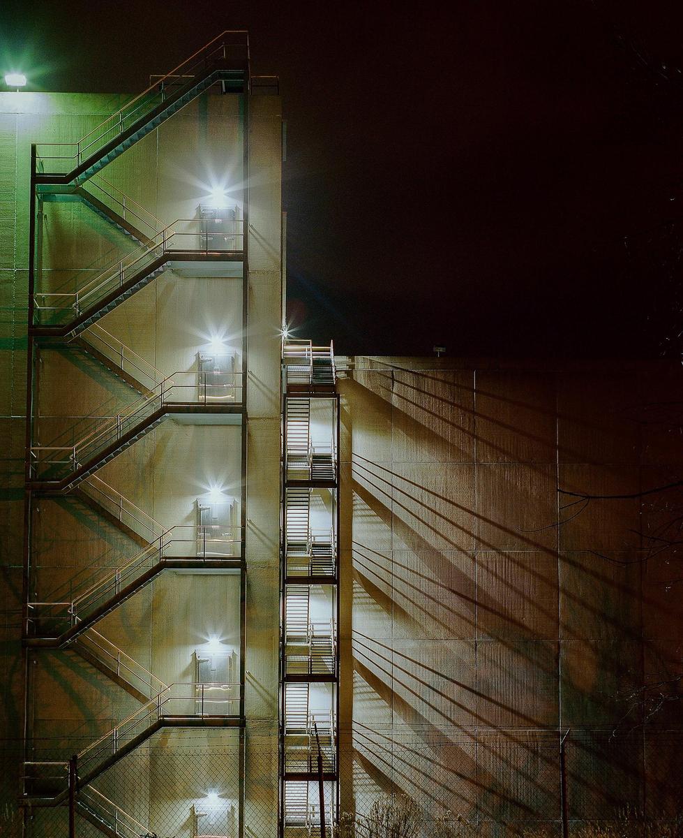Two fire escapes with bright lights cast long shadows on the side of a building at night.