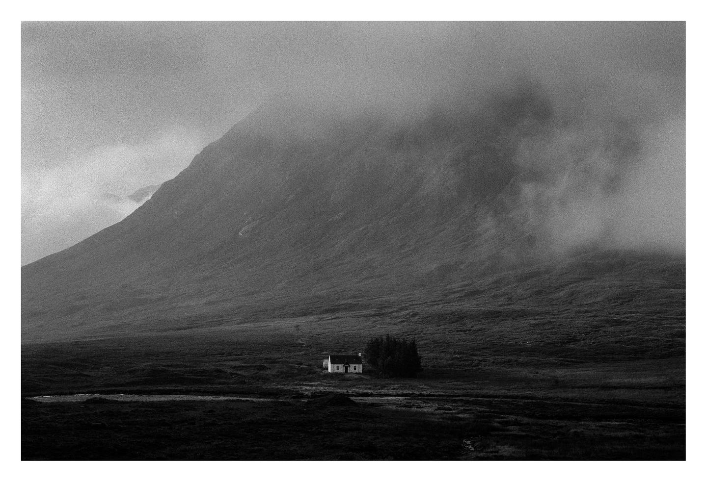 A small house and a cluster of trees sit in a vast, misty valley at the base of a large mountain.