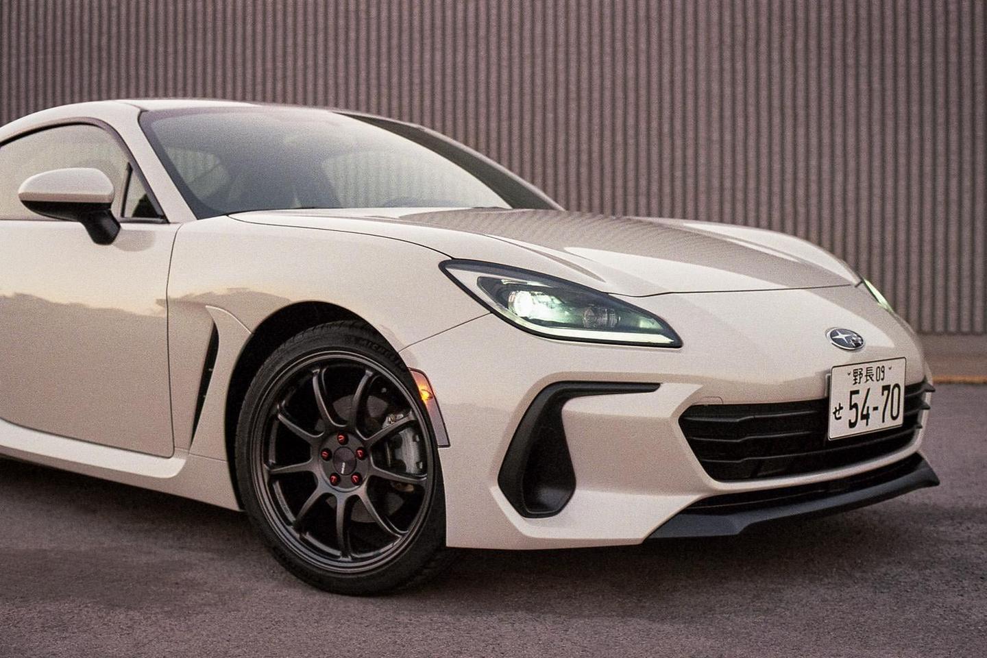 A white sports car with a Japanese license plate is parked on asphalt next to a corrugated metal wall.