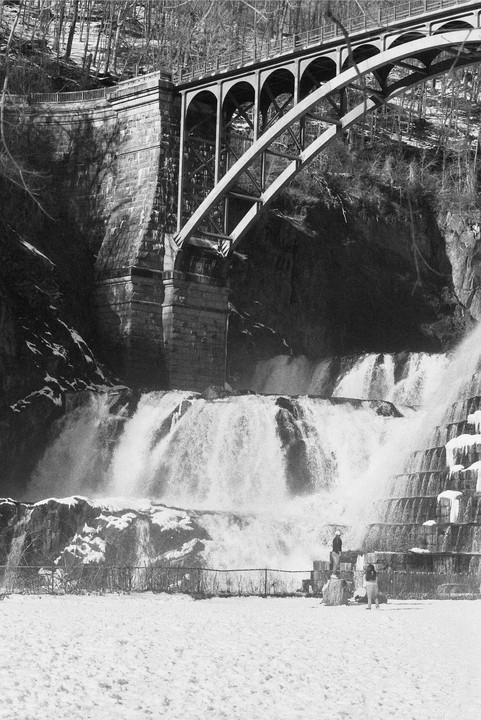A bridge spans over a multi-tiered waterfall with two people standing in the snow at its base.