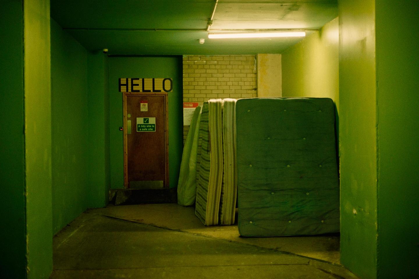 A dark green hallway with a door that has the word "HELLO" above it and several mattresses leaning against a brick wall.