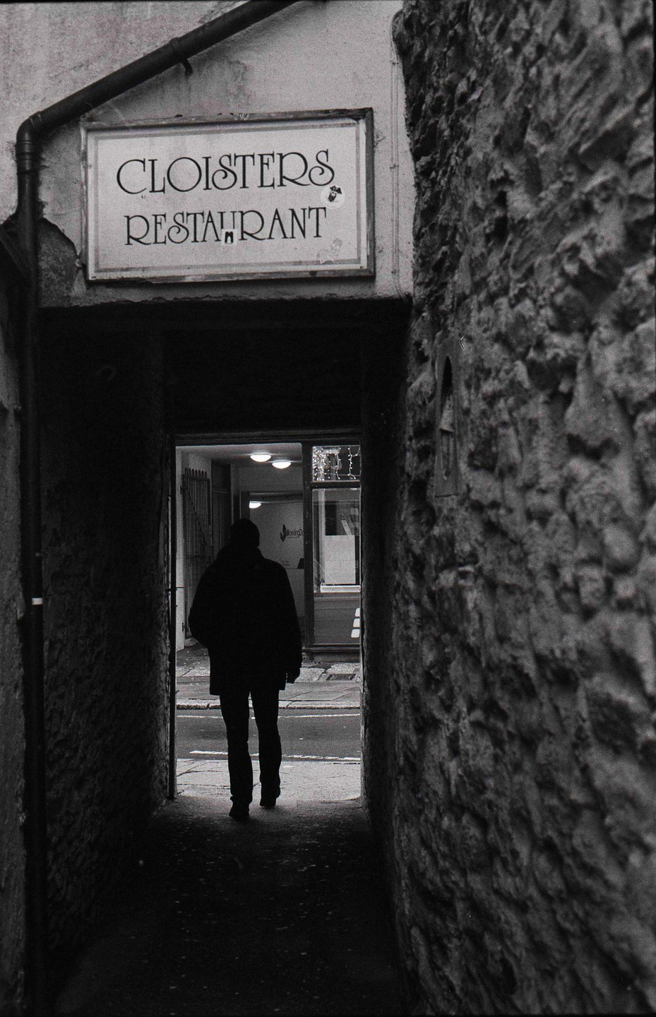 A person walks through a dark stone archway towards a brightly lit street with a restaurant sign above the archway.