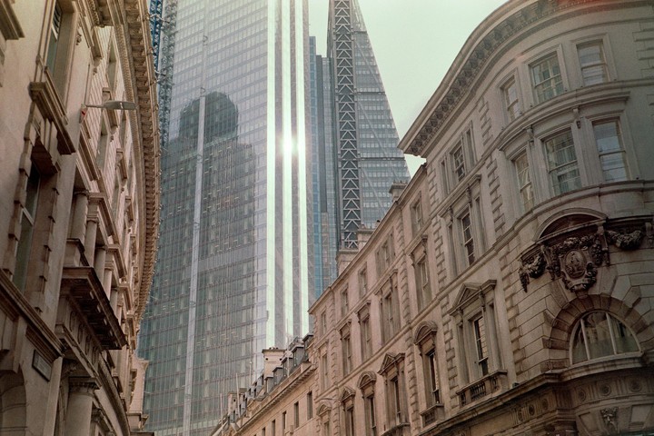 Sunlight reflects off a modern glass skyscraper towering over traditional stone buildings in a city.