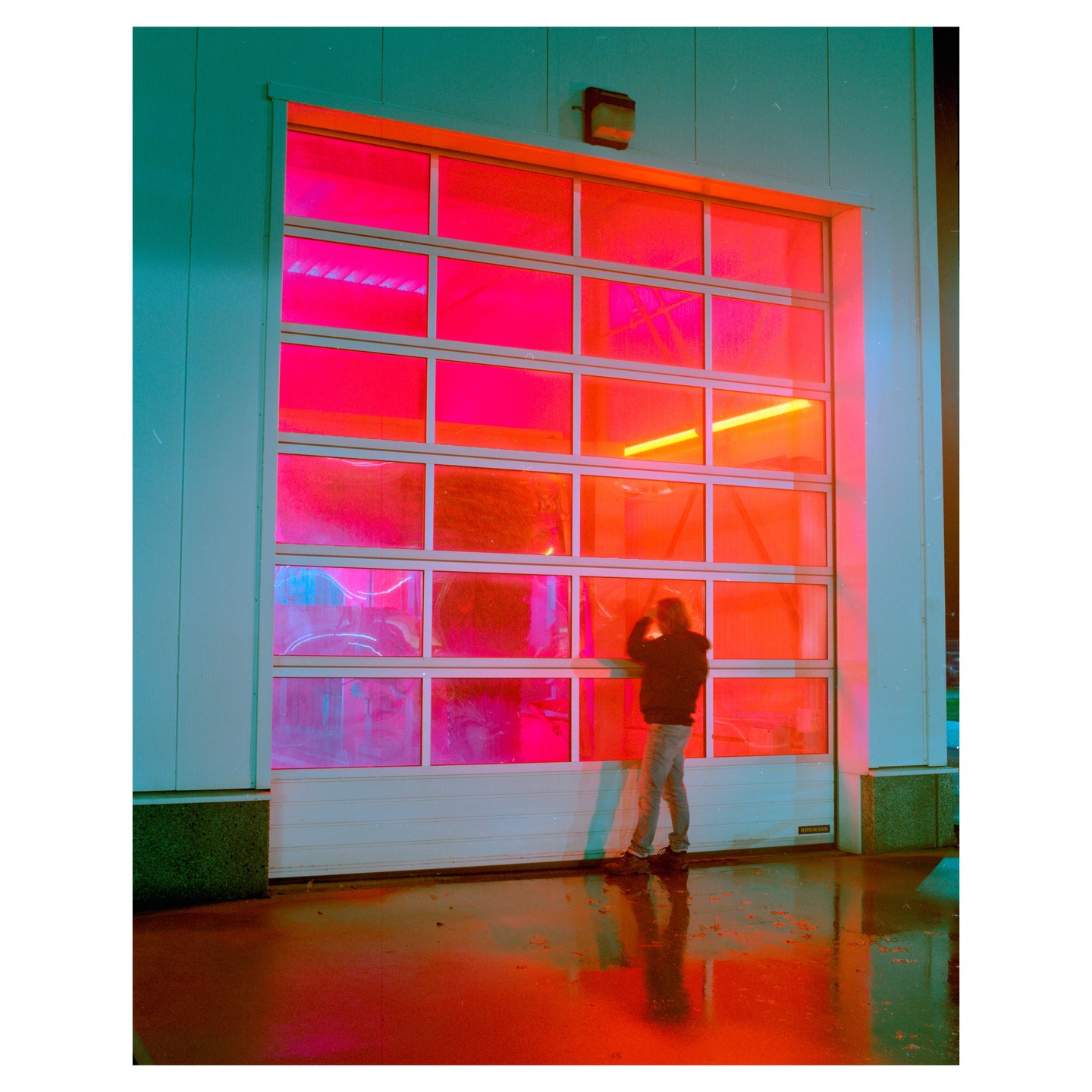 A person stands in front of a garage door with bright pink and orange light shining through its windows, reflecting on the wet ground.