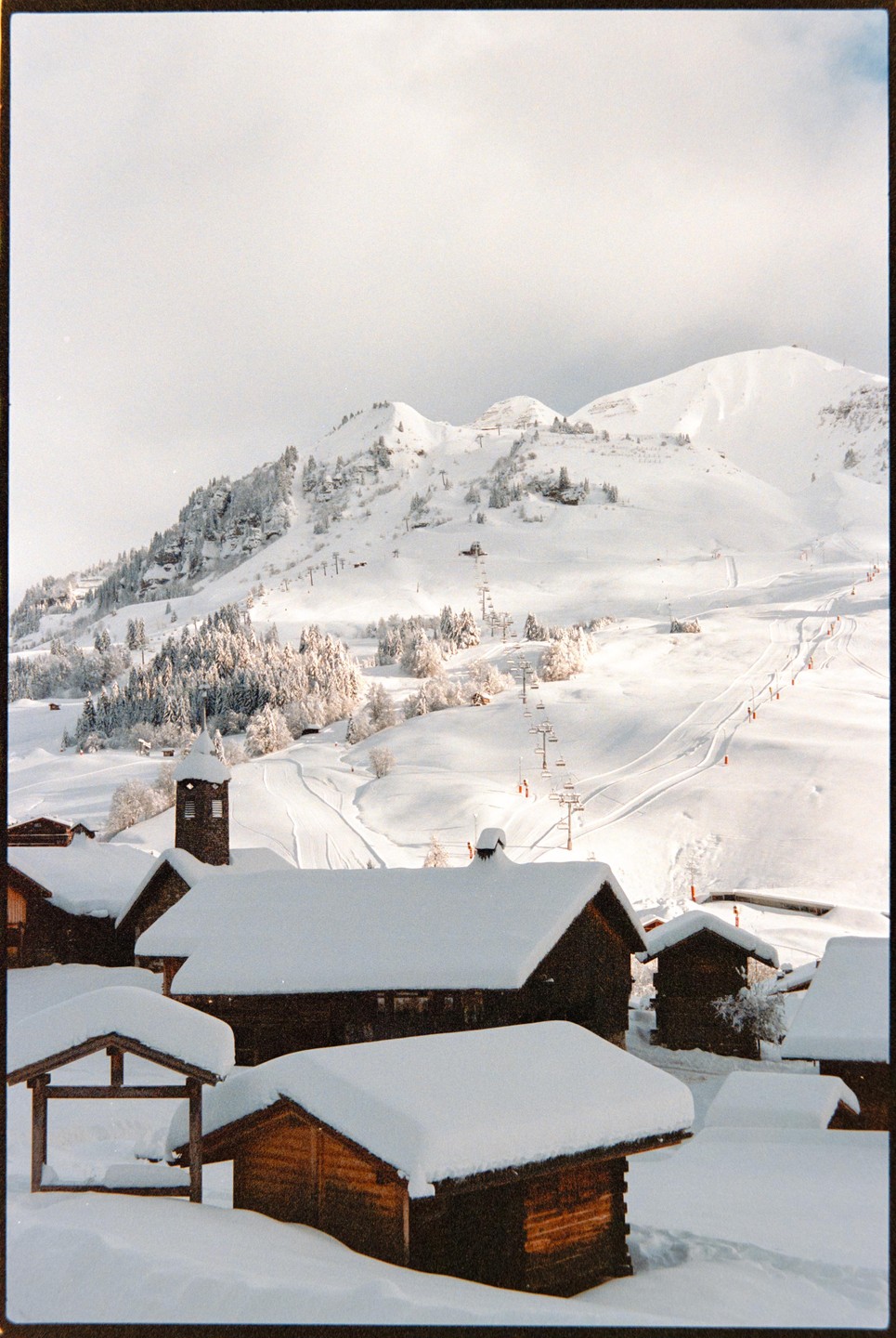 A snow-covered village with wooden buildings sits in the foreground of a mountain landscape with ski slopes and chairlifts.
