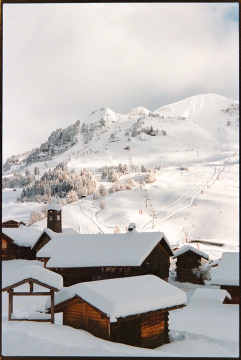 A snow-covered village with wooden buildings sits in the foreground of a mountain landscape with ski slopes and chairlifts.