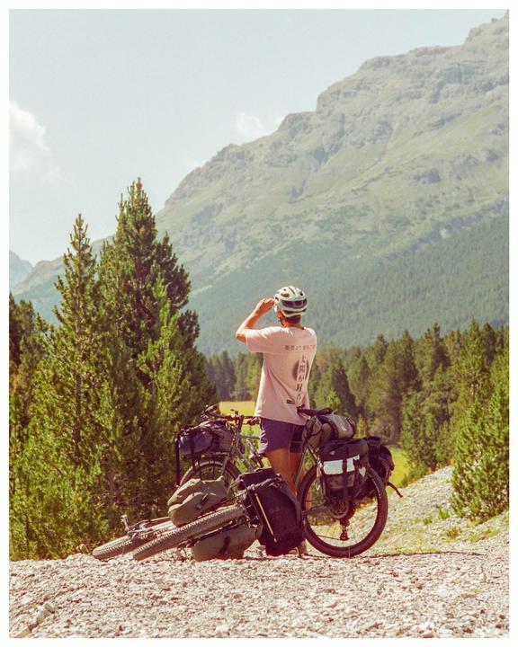 A person wearing a helmet and t-shirt stands next to a loaded bicycle on a gravel path with mountains and trees in the background.