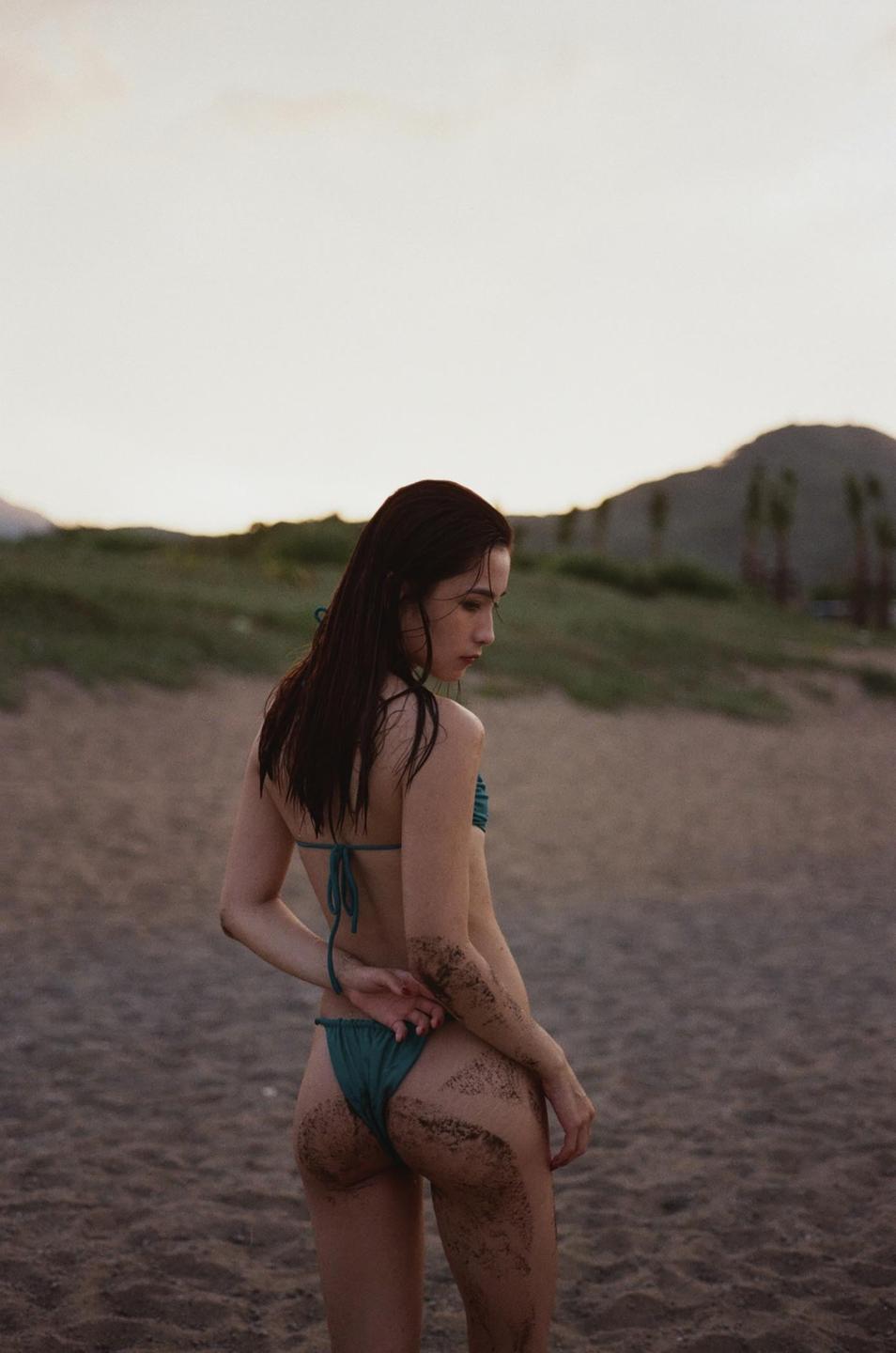 A woman in a bikini stands on a sandy beach with mountains in the background.