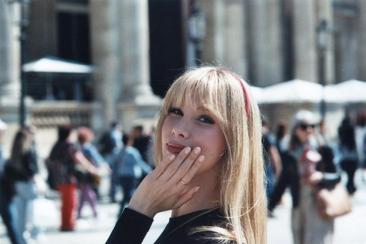 A woman with long blonde hair and a red headband poses for a portrait with her hand near her mouth, with a blurred background of people and architecture.