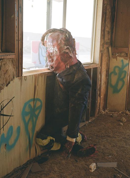 A mannequin with a red, horned head and a dark suit kneels by a window in a dilapidated room with graffiti on the walls.