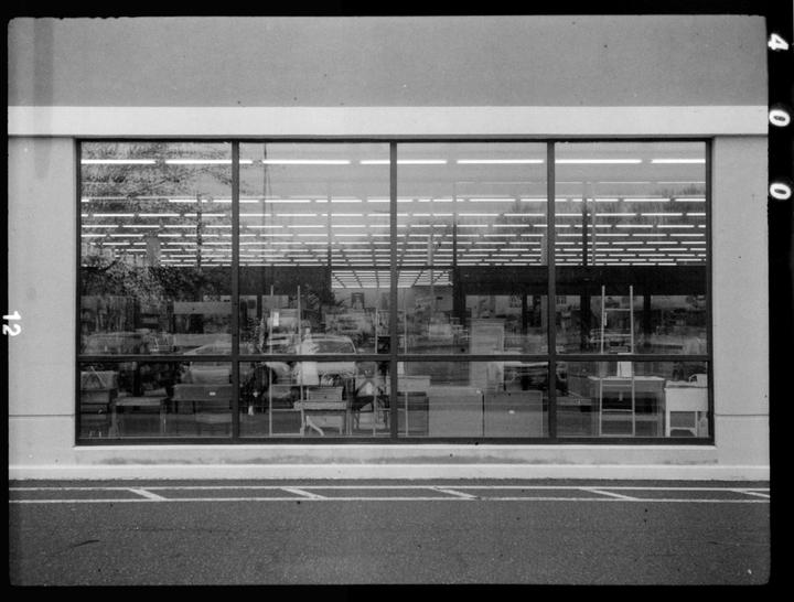 A large storefront window reflects trees and the interior of a store with fluorescent lights.