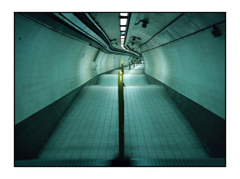 A long, empty underground tunnel with a tiled floor and yellow poles leading into the distance.