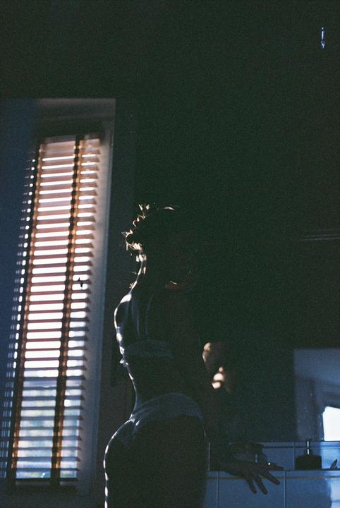 A woman stands in a dark room with light coming through a window with blinds.