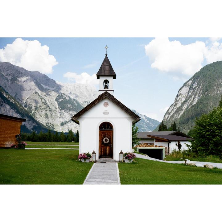 A small white chapel with a brown roof and bell tower stands in a green field with mountains in the background.
