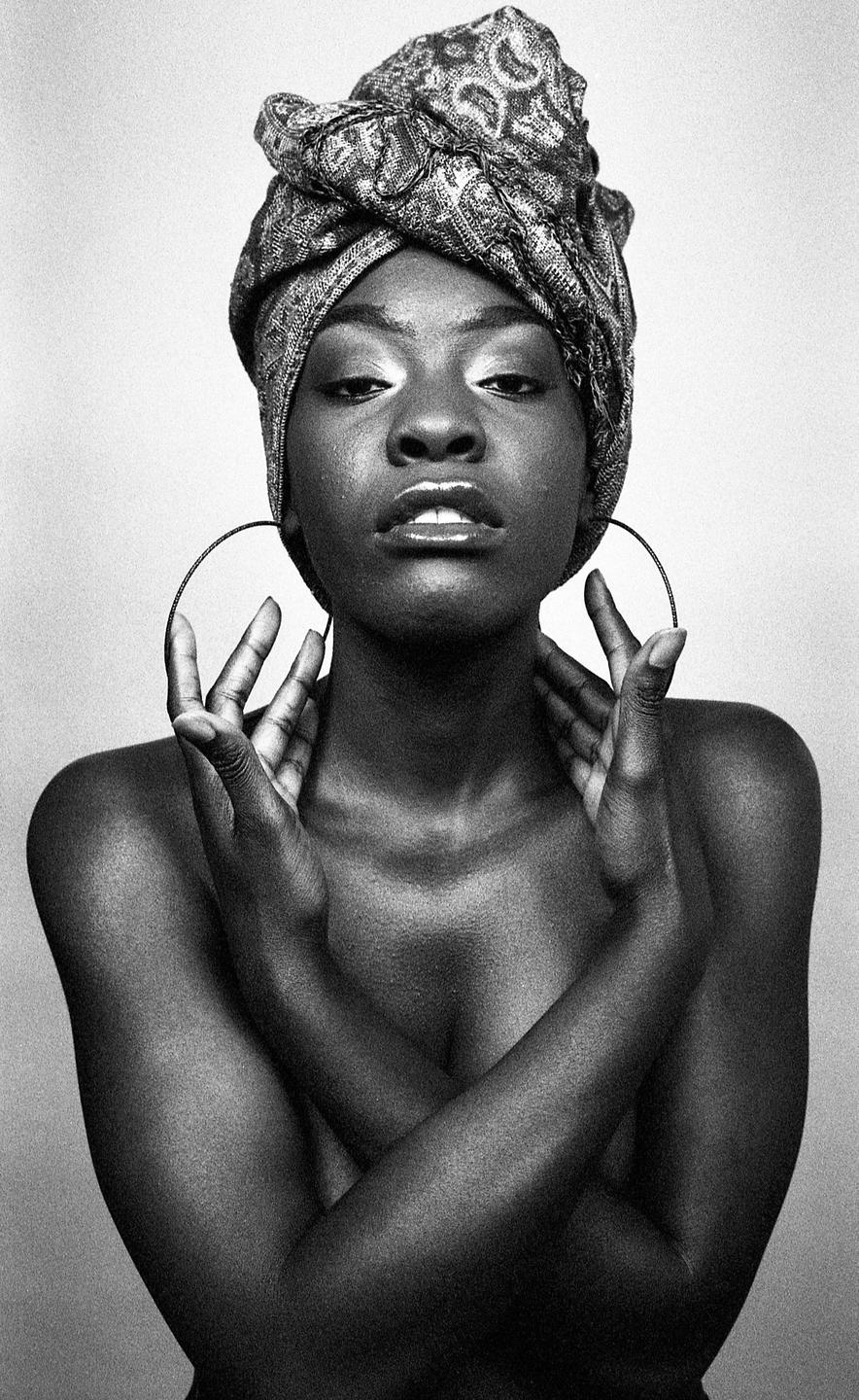 A woman with a patterned head wrap and large hoop earrings poses with her arms crossed over her chest and hands near her face.