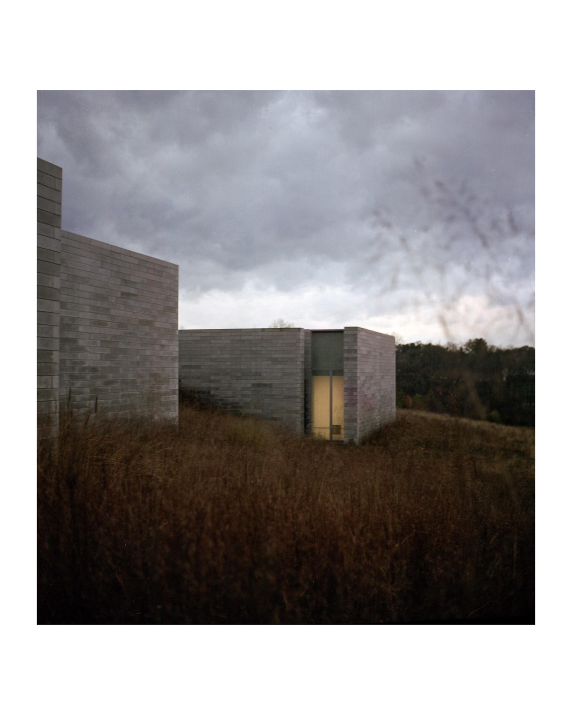 Two modern buildings with light coming from a window are surrounded by tall, dry grass under a cloudy sky.