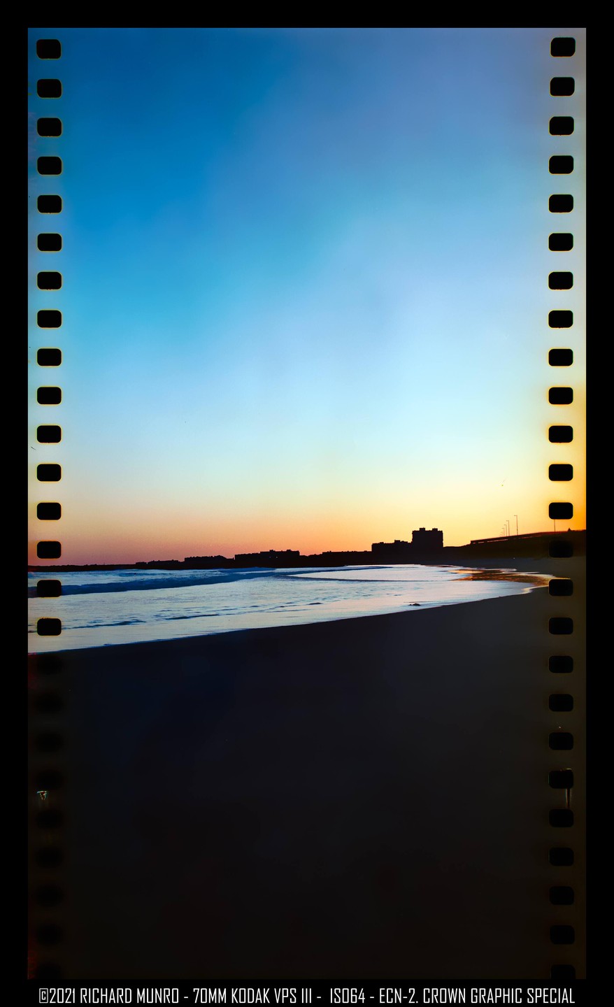 A beach at sunset shows waves crashing on the shore with buildings silhouetted in the distance.