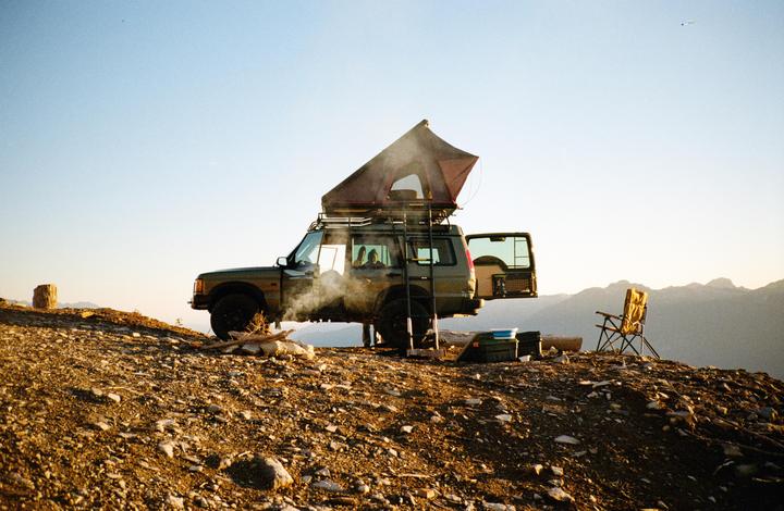 A vehicle with a rooftop tent is parked on a rocky hill with mountains in the background.