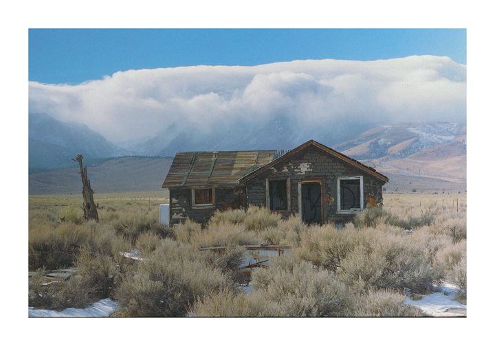 An abandoned shack sits in a field of dry brush with mountains and a cloudy sky in the background.