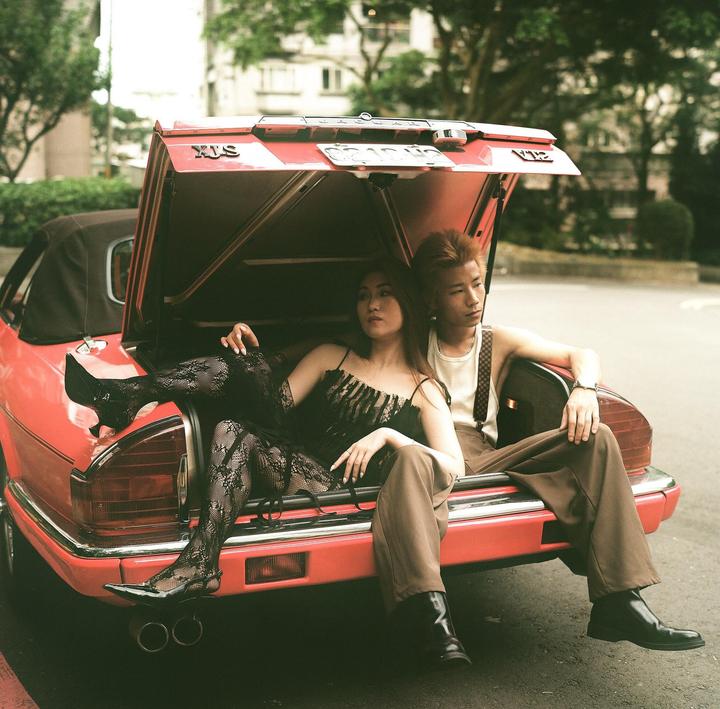 A couple sits in the open trunk of a red convertible car on a street.