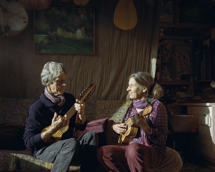 A man and a woman are sitting on a sofa playing string instruments in a sunlit room.