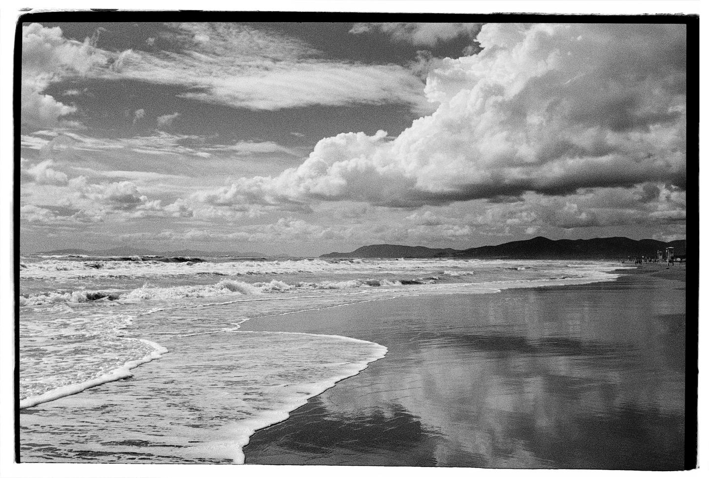 Waves crash on a beach under a cloudy sky with mountains in the distance.