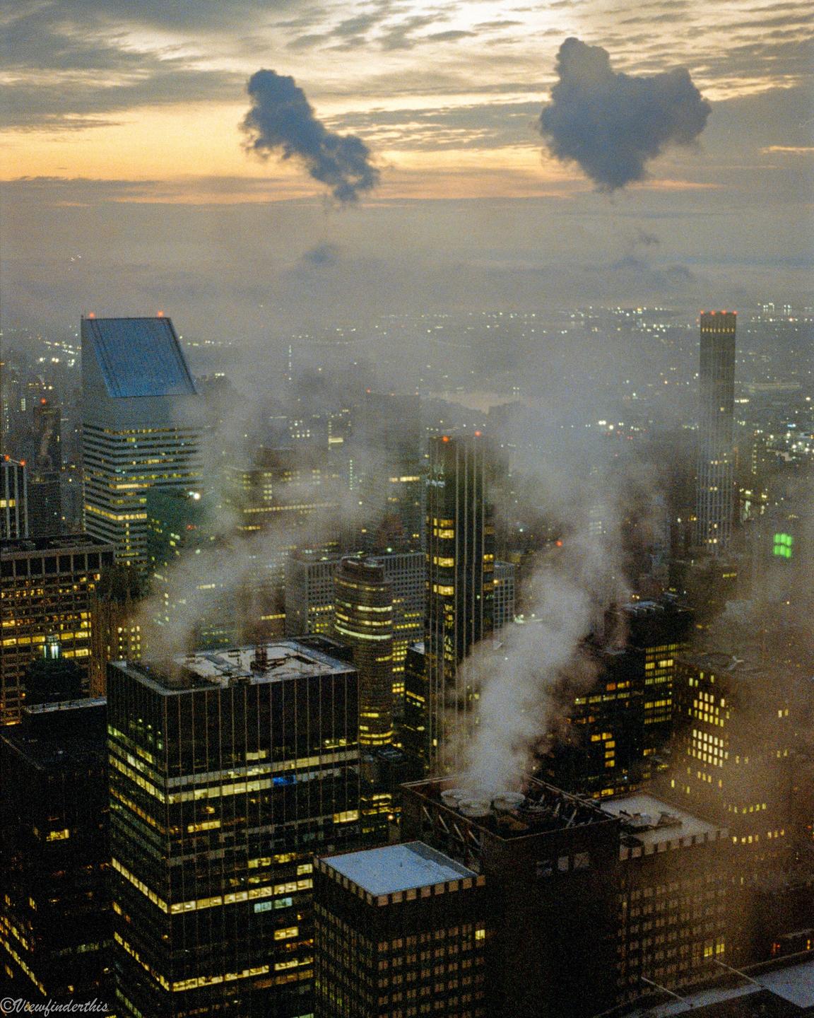 An aerial view of a city at dusk shows illuminated buildings and steam rising from rooftops, with a hazy sky above.