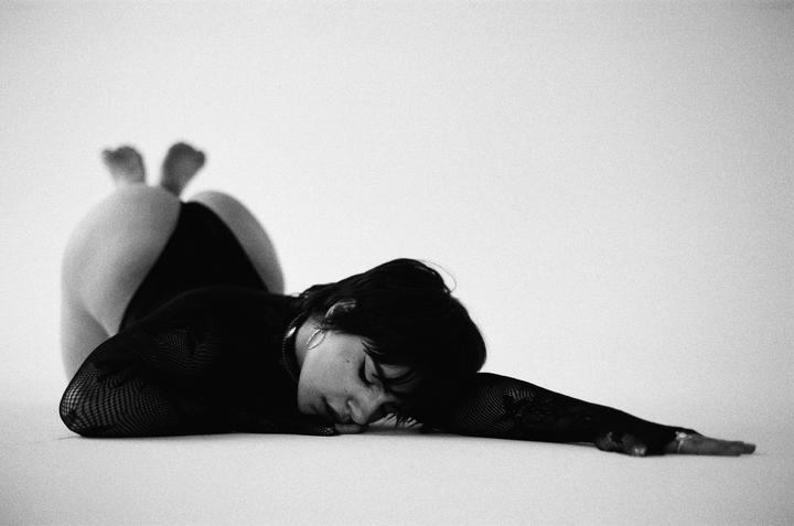 A woman in a black mesh top is lying on her stomach with her head resting on her arm, and her legs are bent with her feet pointing upwards.
