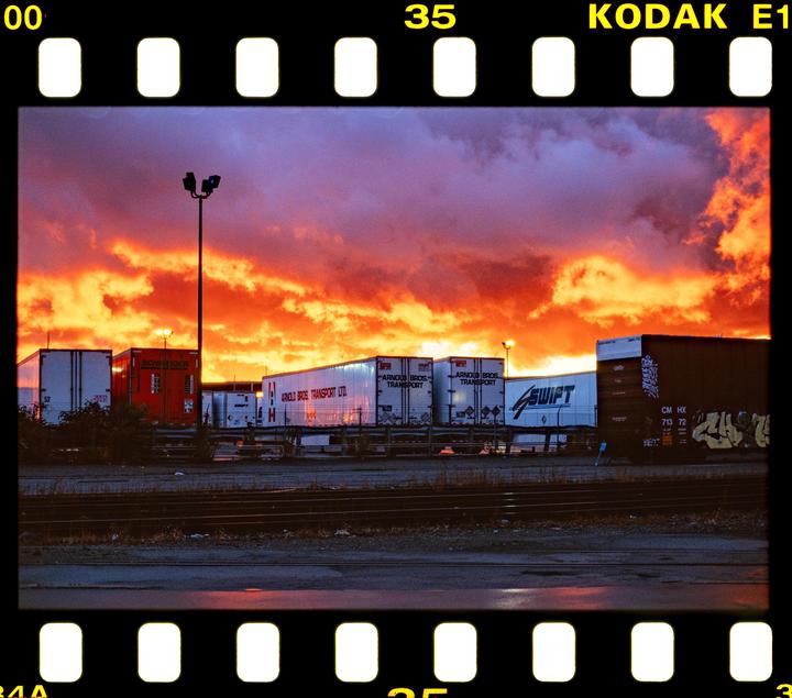 A dramatic sunset with orange and red clouds illuminates a row of parked semi-trailers and a street light.