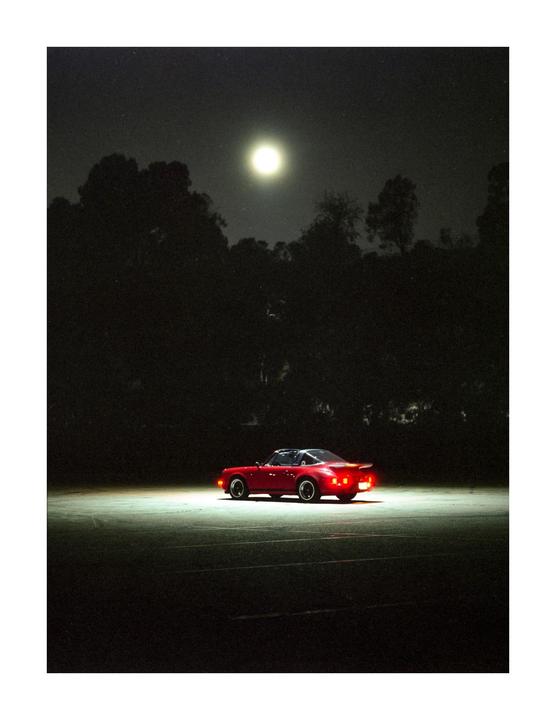 A red sports car is parked on an empty lot under a bright full moon at night.