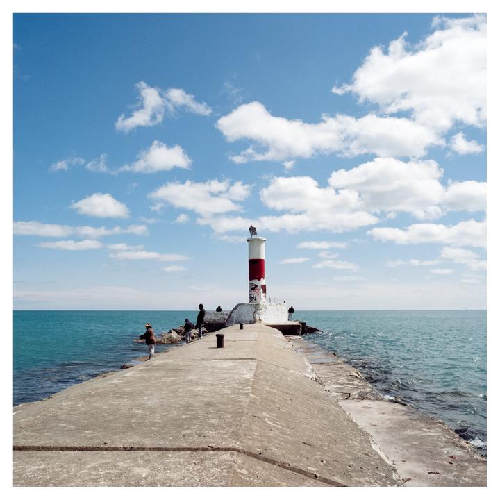 A red and white lighthouse stands at the end of a concrete pier with people gathered around it under a blue sky with white clouds.