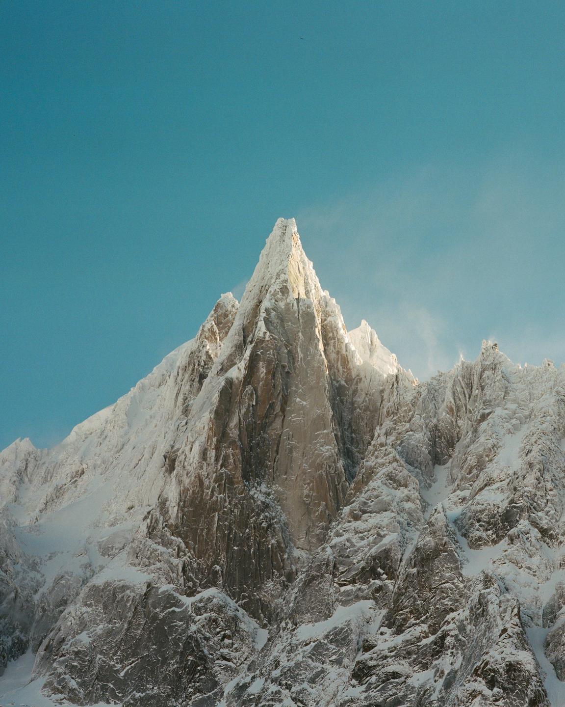 A snow-covered mountain peak rises sharply against a clear blue sky.