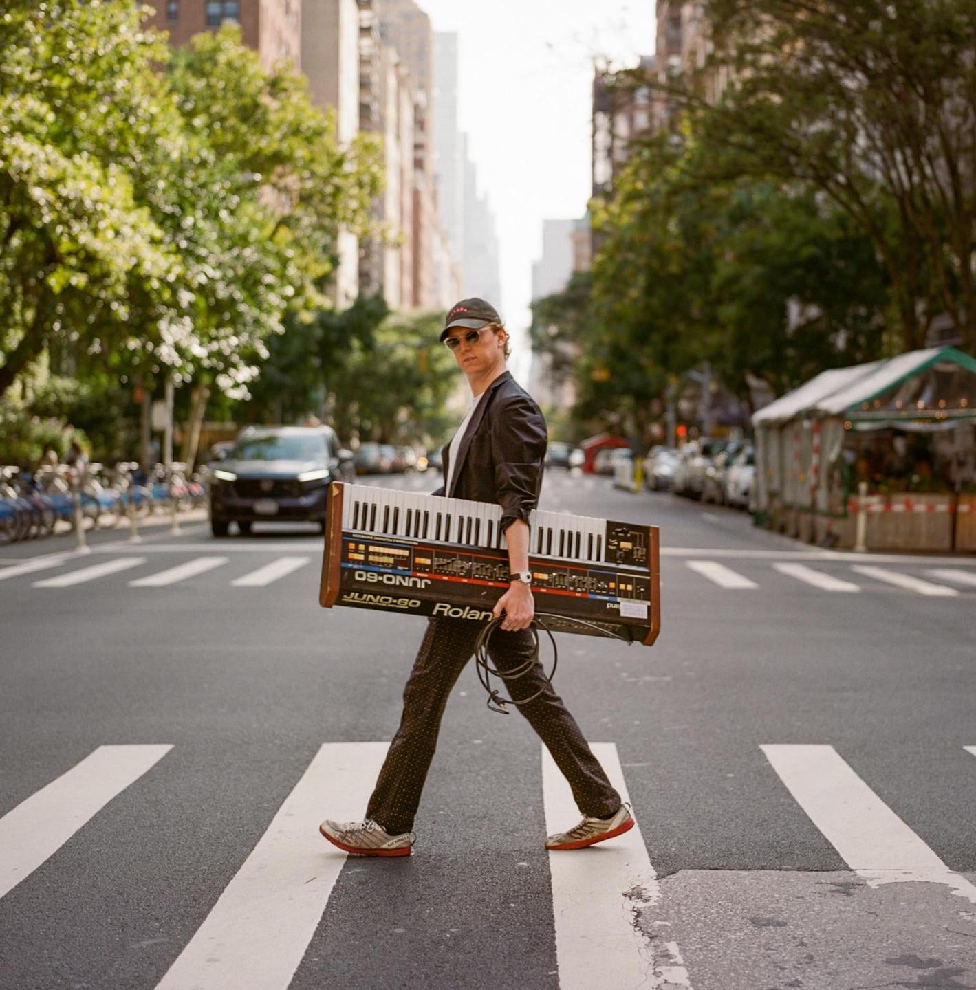 A man carrying a synthesizer crosses a street in a city.