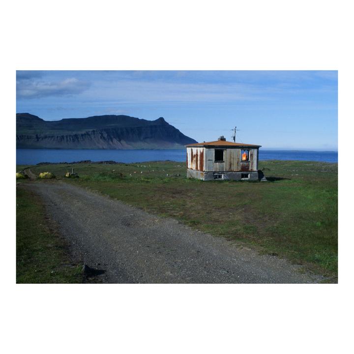 A small, weathered building with a glowing window sits on a grassy shore next to a body of water, with mountains in the distance.