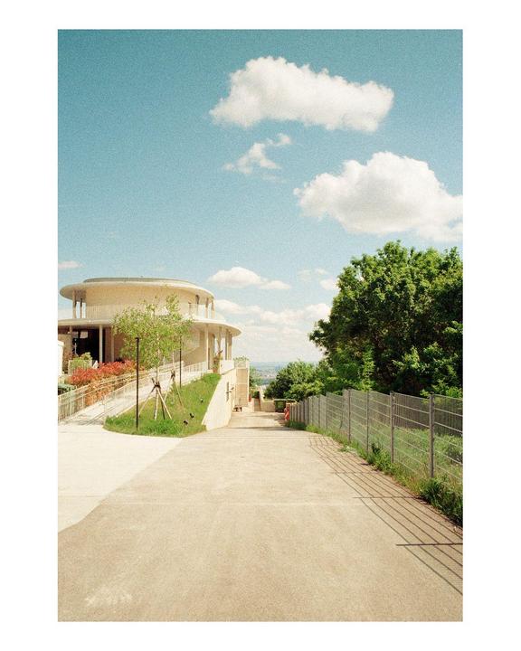 A modern round building with balconies and a fence next to a paved path under a blue sky with clouds.