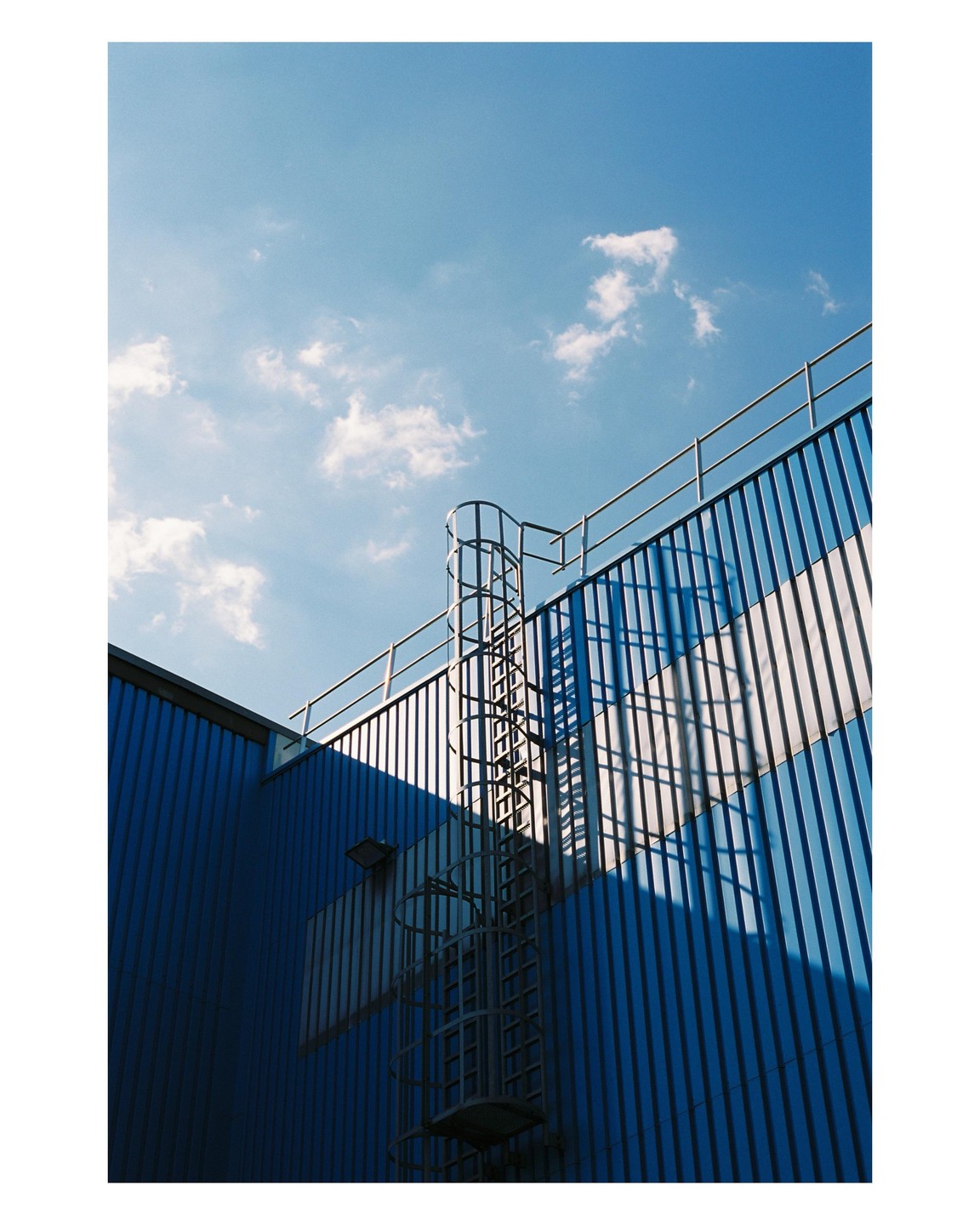 A blue corrugated metal building with a caged ladder and railing is shown under a blue sky with white clouds.