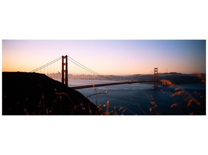 The Golden Gate Bridge spans a body of water with a city skyline in the distance under a colorful sky.