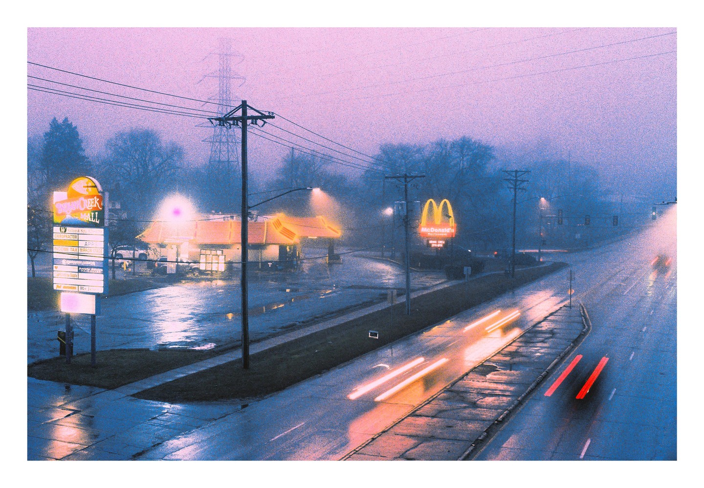 A McDonald's restaurant and a shopping mall sign are visible through a thick fog at dusk, with light trails from cars on a wet road.