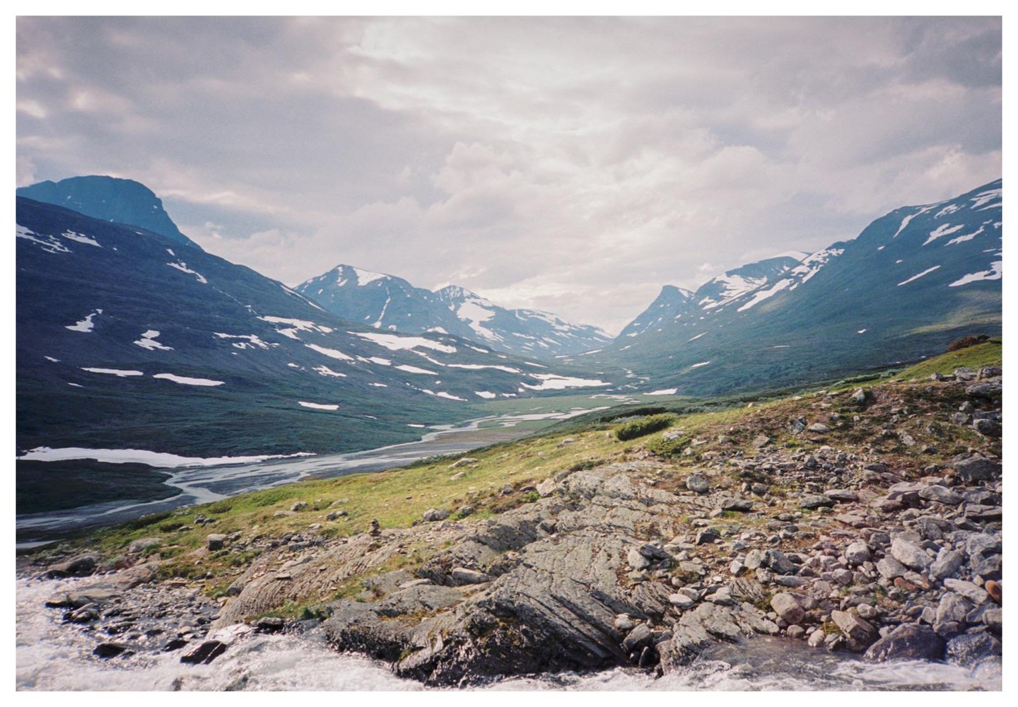 A wide valley with a river running through it is surrounded by mountains with patches of snow under a cloudy sky.